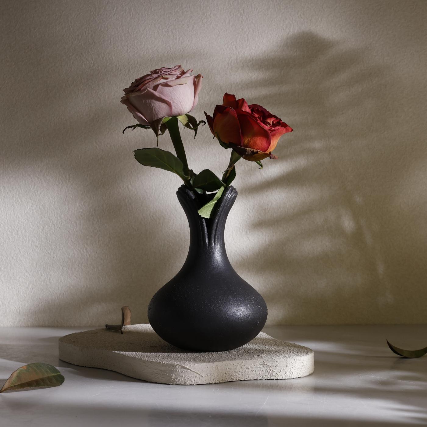 Lifestyle shot of the BosilunLife sculptural black flower vase in recycled eco-clay. Resting on a textured stone slab, the matte black vase holds a dusty pink and a deep red rose. Dramatic botanical shadows fall across the warm beige background, showcasing a moody, organic modern, and wabi-sabi home décor styling.