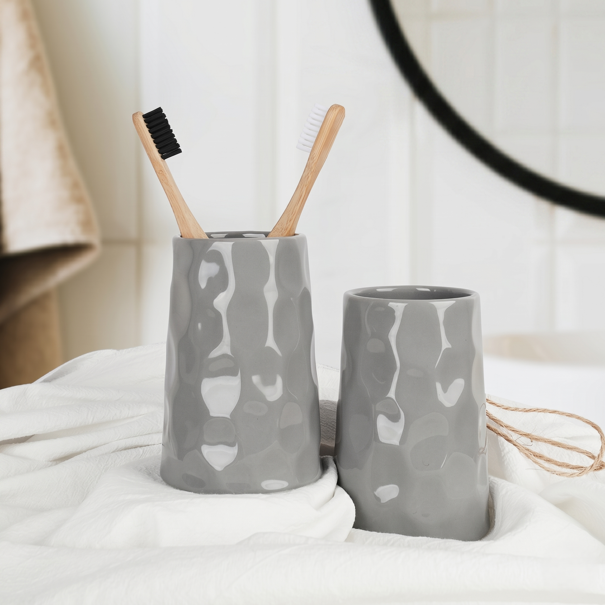 Close-up lifestyle shot of the BosilunLife Bring Nature Inside grey ceramic toothbrush holder and matching tumbler. The glossy, organic rippled texture of the recycled eco-clay catches the light elegantly against a draped white towel. Two sustainable bamboo toothbrushes rest inside, perfectly capturing a calming, spa-inspired bathroom aesthetic.