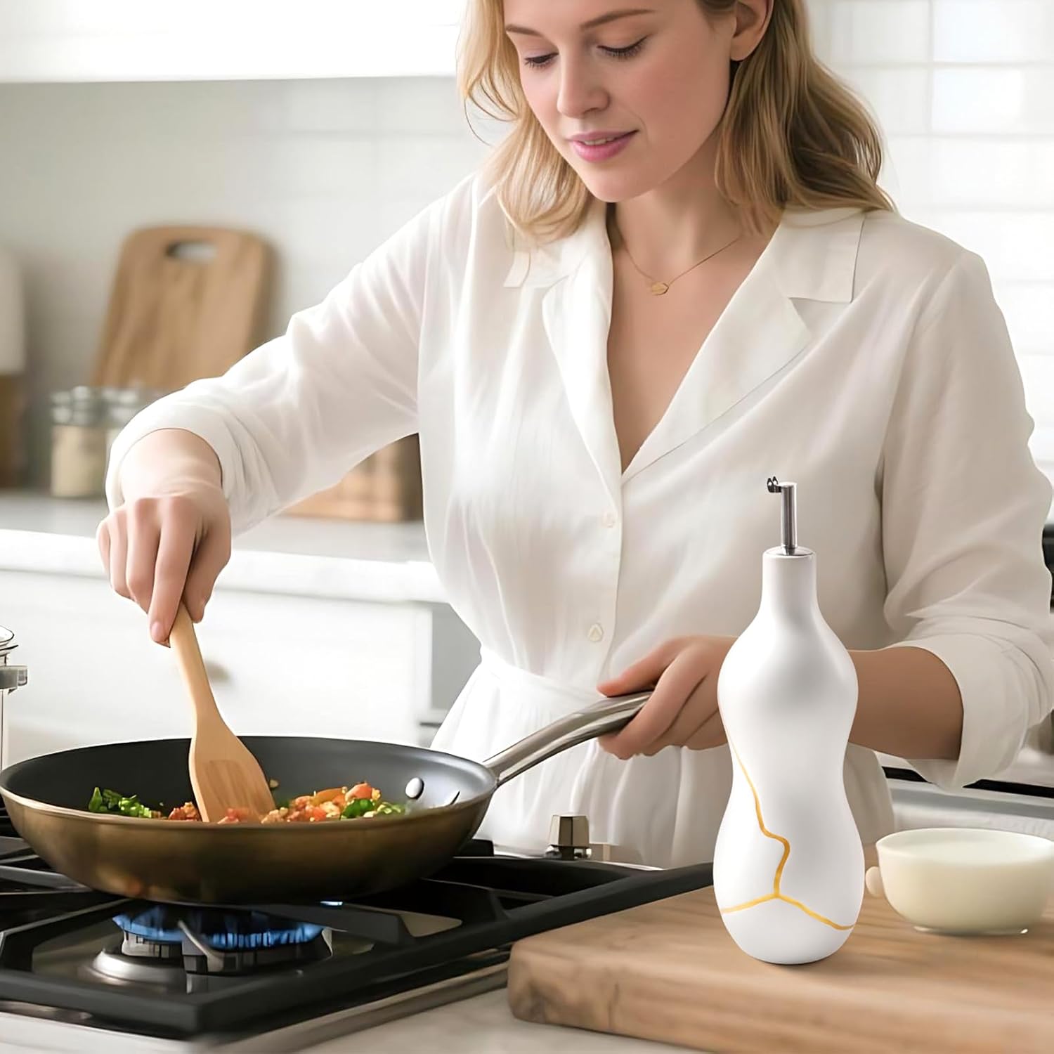 A lifestyle photograph of the BosilunLife 22 oz ceramic oil cruet in matte white, featuring a striking Kintsugi-inspired gold vein pattern and a stainless steel pourer spout. The sculptural bottle rests on a light-colored wooden cutting board on a kitchen countertop. Surrounding it are fresh green basil leaves and a raw brown egg, suggesting healthy meal preparation. The background shows a gently blurred, bright modern kitchen with stone surfaces and wooden utensils, illustrating an elegant cooking enviornm