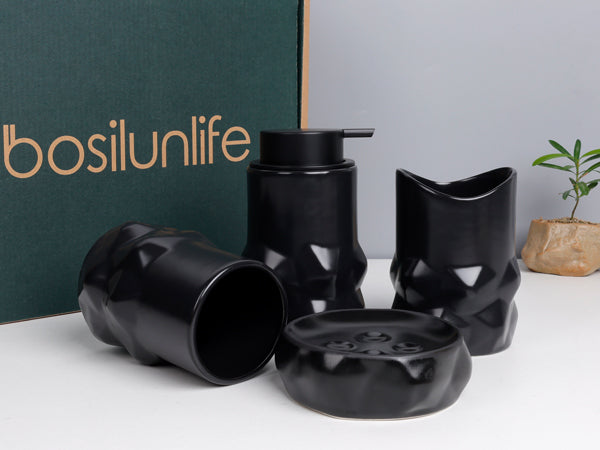 Premium unboxing shot of the BosilunLife 4-piece diamond-shaped bathroom accessory set in matte black recycled eco-clay. Arranged in front of a dark green branded BosilunLife gift box, the sculptural, faceted collection features a manual liquid soap dispenser, a round soap dish, an upright curved-rim tumbler, and a flat-rimmed tumbler resting on its side. A small potted plant adds a touch of organic modern styling, highlighting an elegant, ready-to-gift presentation.