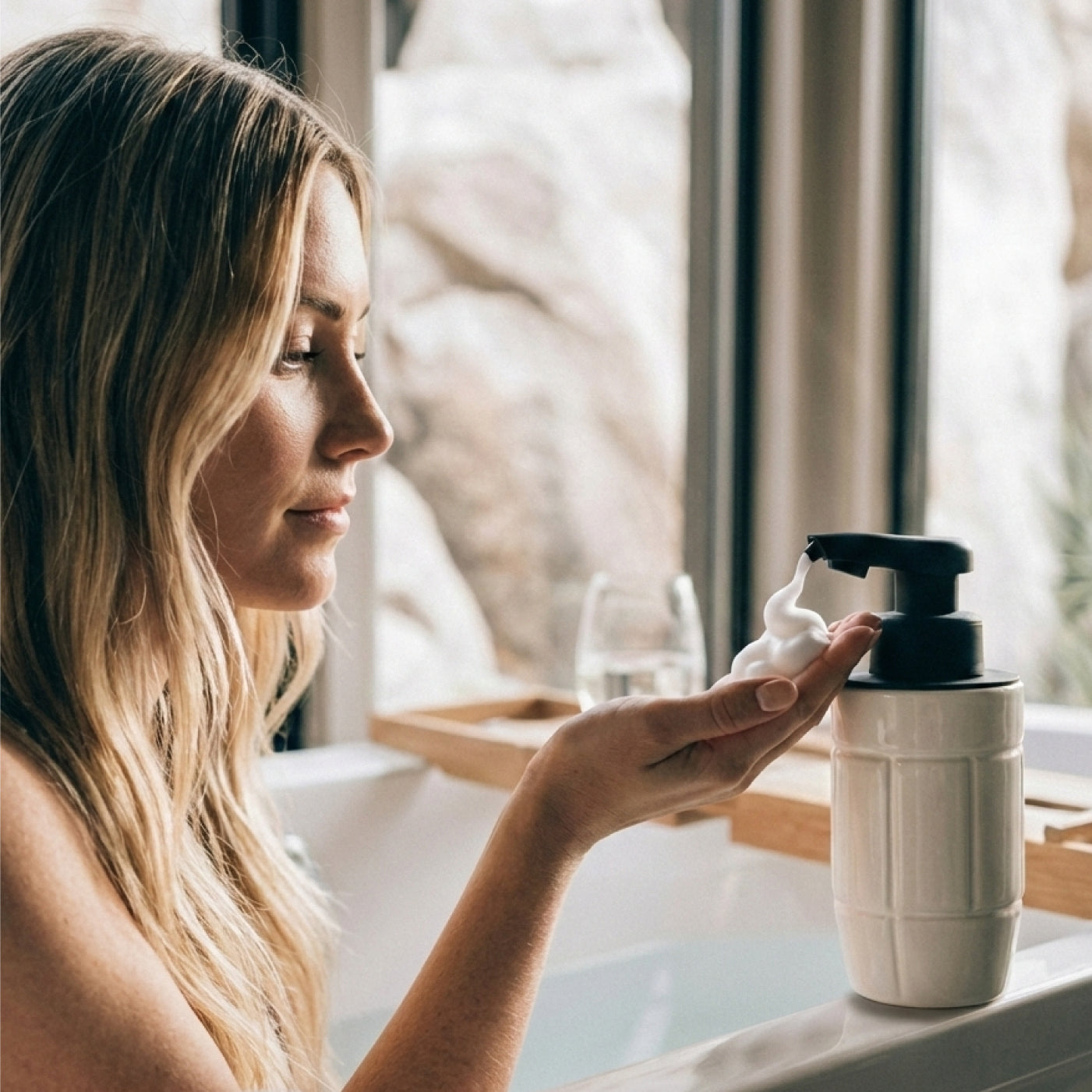 Lifestyle shot of a woman using the BosilunLife Eco-Brutalism automatic foaming soap dispenser whilst relaxing by a bath. The off-white recycled eco-clay dispenser, featuring an embossed grid pattern, sits on the edge of the tub. The touchless black sensor pump automatically dispenses rich white foam into her hand, set against a large window framing a rugged rock landscape for a serene, spa-inspired aesthetic.