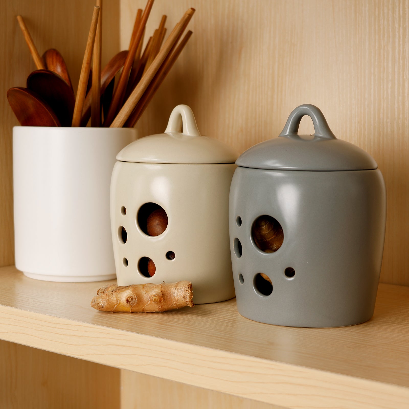 Lifestyle styling shot of two BosilunLife ceramic storage jars in ecru and cool grey recycled eco-clay, sitting on a light wooden pantry shelf. The breathable, perforated keepers are filled with fresh ginger and shallots, with a piece of raw ginger resting in front. Styled beside a white ceramic utensil holder filled with wooden spoons and chopsticks, the image showcases versatile, odour-resistant root vegetable storage and elegant kitchen organisation.