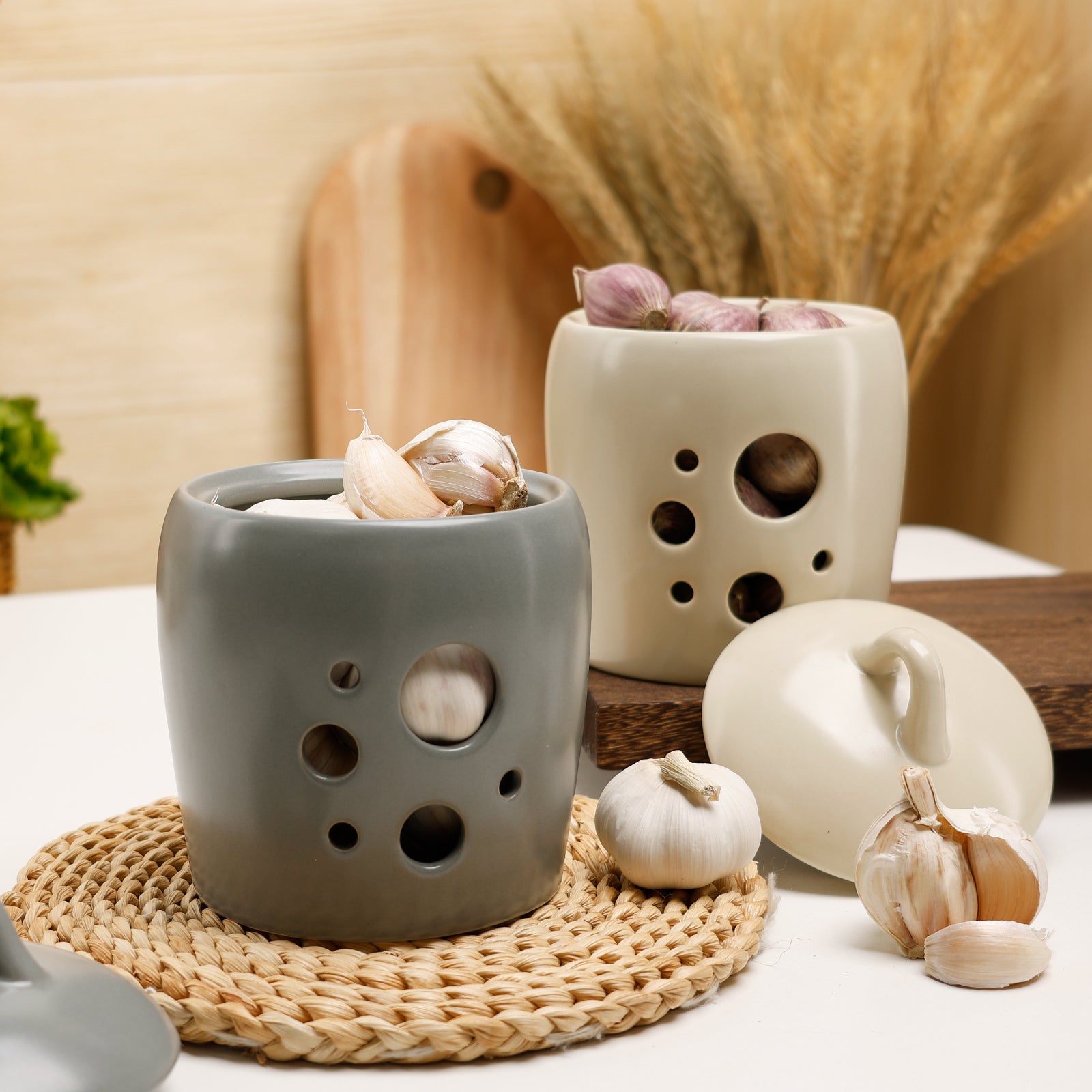 Lifestyle composition of two open BosilunLife ceramic garlic keepers in cool grey and smooth ecru recycled eco-clay. Styled in a warm, rustic kitchen setting, the grey jar sits on a woven rattan trivet overflowing with white garlic cloves, whilst the ecru jar rests on a dark wooden board filled with purple garlic. Surrounded by fresh bulbs, dried wheat, and a wooden chopping board, the image highlights high-capacity, odour-resistant worktop storage with a cottagecore farmhouse aesthetic.