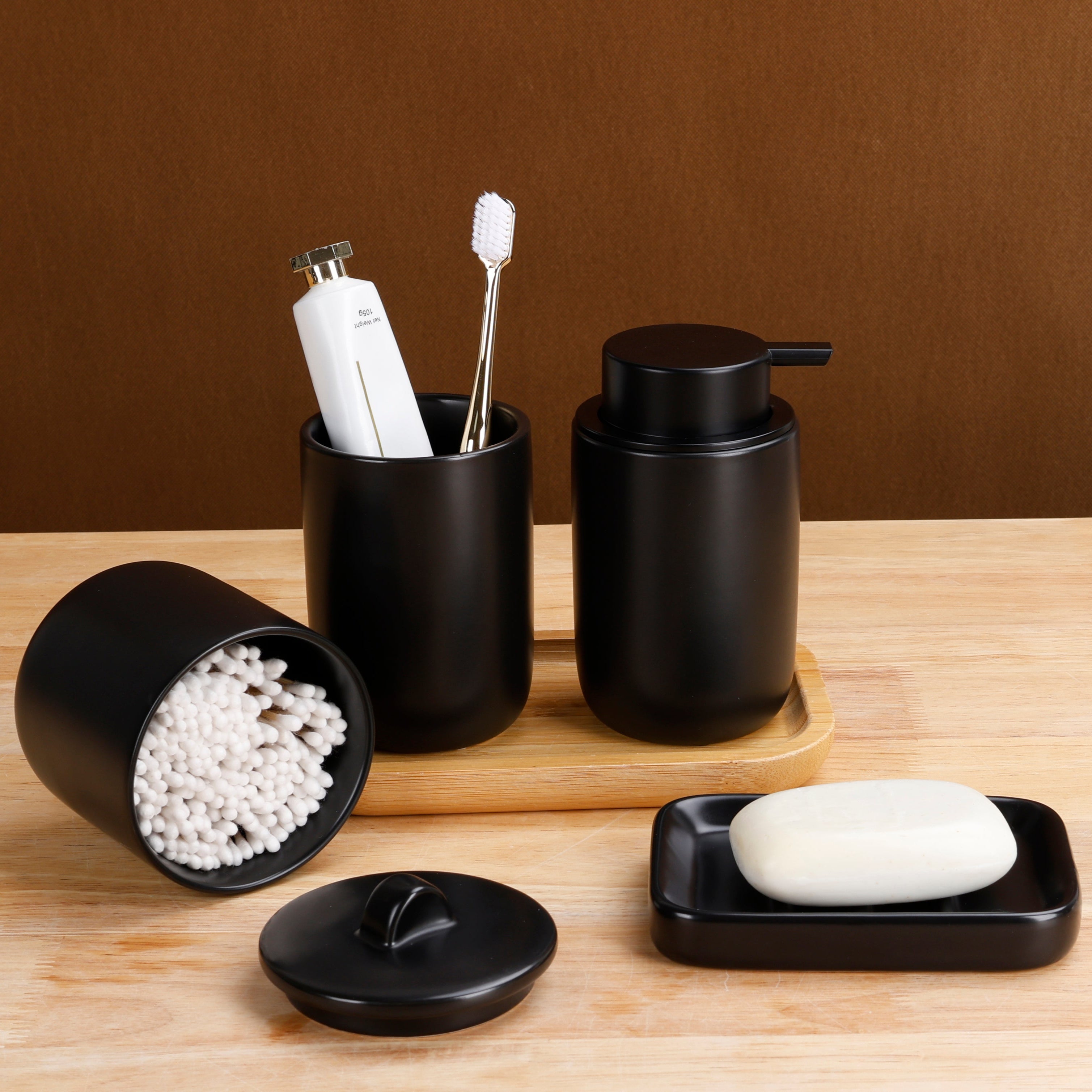 Functional lifestyle shot of the BosilunLife 4-piece matte black bathroom accessory set in recycled eco-clay. Styled on a warm wooden surface, the collection features a manual soap dispenser and a toothbrush holder resting on a wooden tray. In the foreground, a rectangular soap dish holds a bar of soap, whilst an open apothecary jar lies on its side to display stored cotton buds, highlighting practical and elegant bathroom organisation.