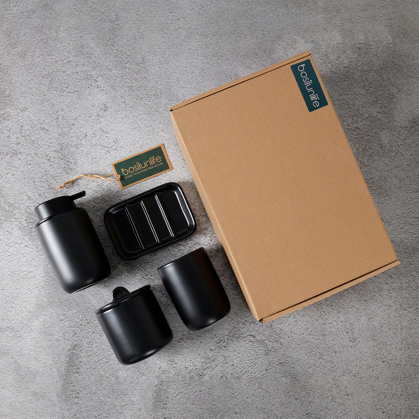 Flat lay unboxing shot of the BosilunLife 4-piece matte black bathroom accessory set in recycled eco-clay. The minimalist collection, featuring a manual soap dispenser, ribbed soap dish, lidded apothecary jar, and tumbler, is arranged on a textured grey concrete surface next to its sustainable kraft cardboard gift box and branded tag, highlighting an eco-luxury gifting experience.
