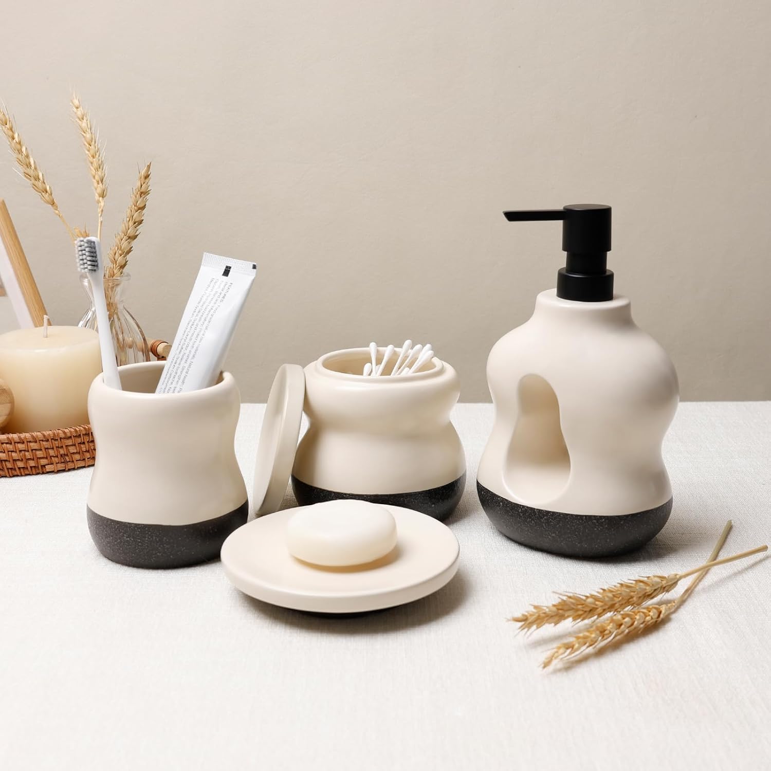 Lifestyle shot of the BosilunLife Pebblebase 4-piece bathroom accessory set in recycled eco-clay. Styled on a light textured cloth alongside dried wheat, the collection features a signature two-tone design with a smooth ecru upper and a dark, speckled pebble base. The set includes a sculptural liquid soap dispenser with a black manual pump, a toothbrush holder, an open lidded jar displaying cotton buds, and a round soap dish holding a white bar of soap, evoking a serene, earthy aesthetic.