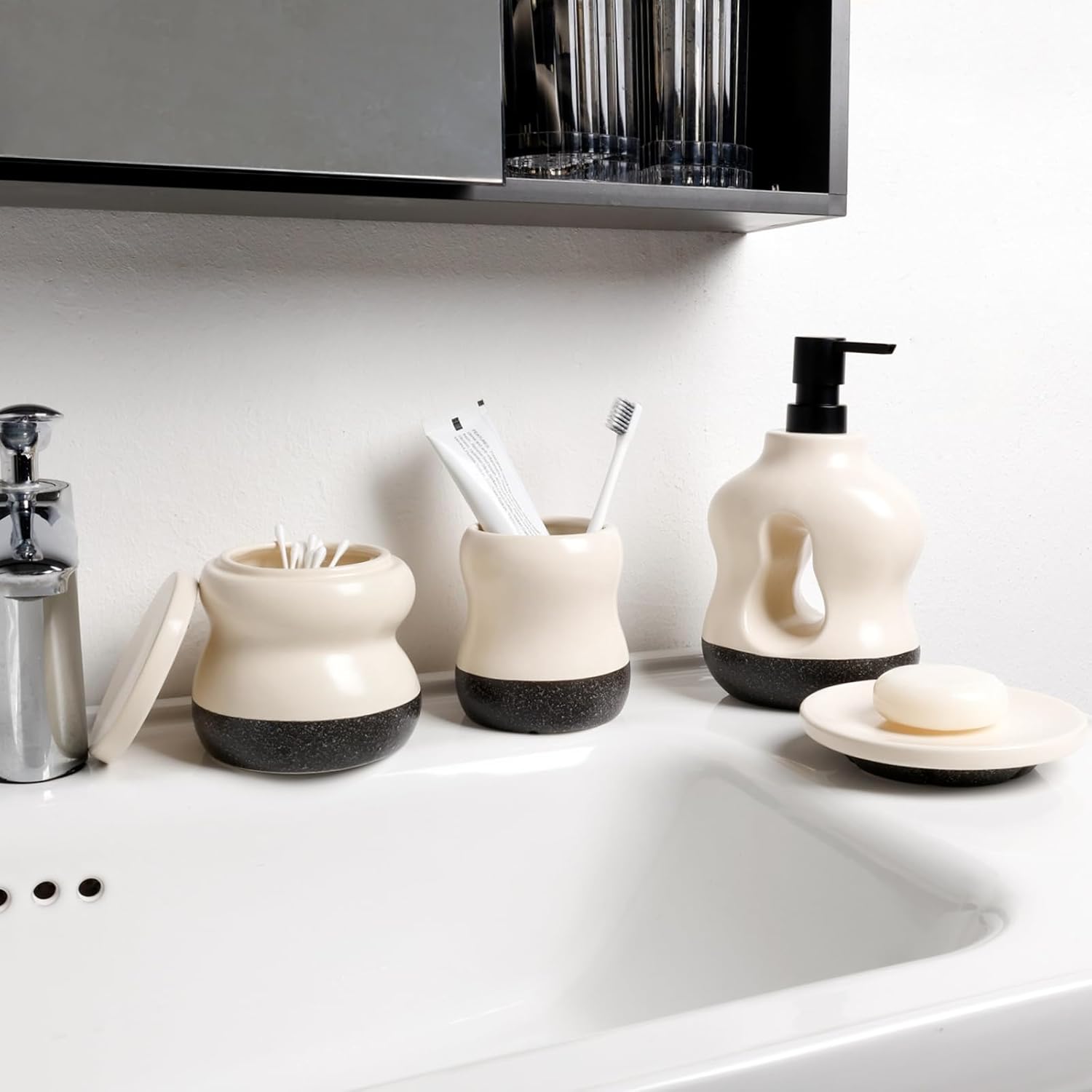 In-situ lifestyle shot of the BosilunLife Pebblebase 4-piece bathroom accessory set in recycled eco-clay, styled on a contemporary white washbasin. The collection features a signature two-tone smooth ecru and dark speckled design, including an open lidded jar displaying cotton buds, a toothbrush holder, a sculptural liquid soap dispenser with a matte black pump, and a round soap dish. Framed by a sleek chrome tap and a black mirrored cabinet, the image showcases elegant, clutter-free washroom organisation.