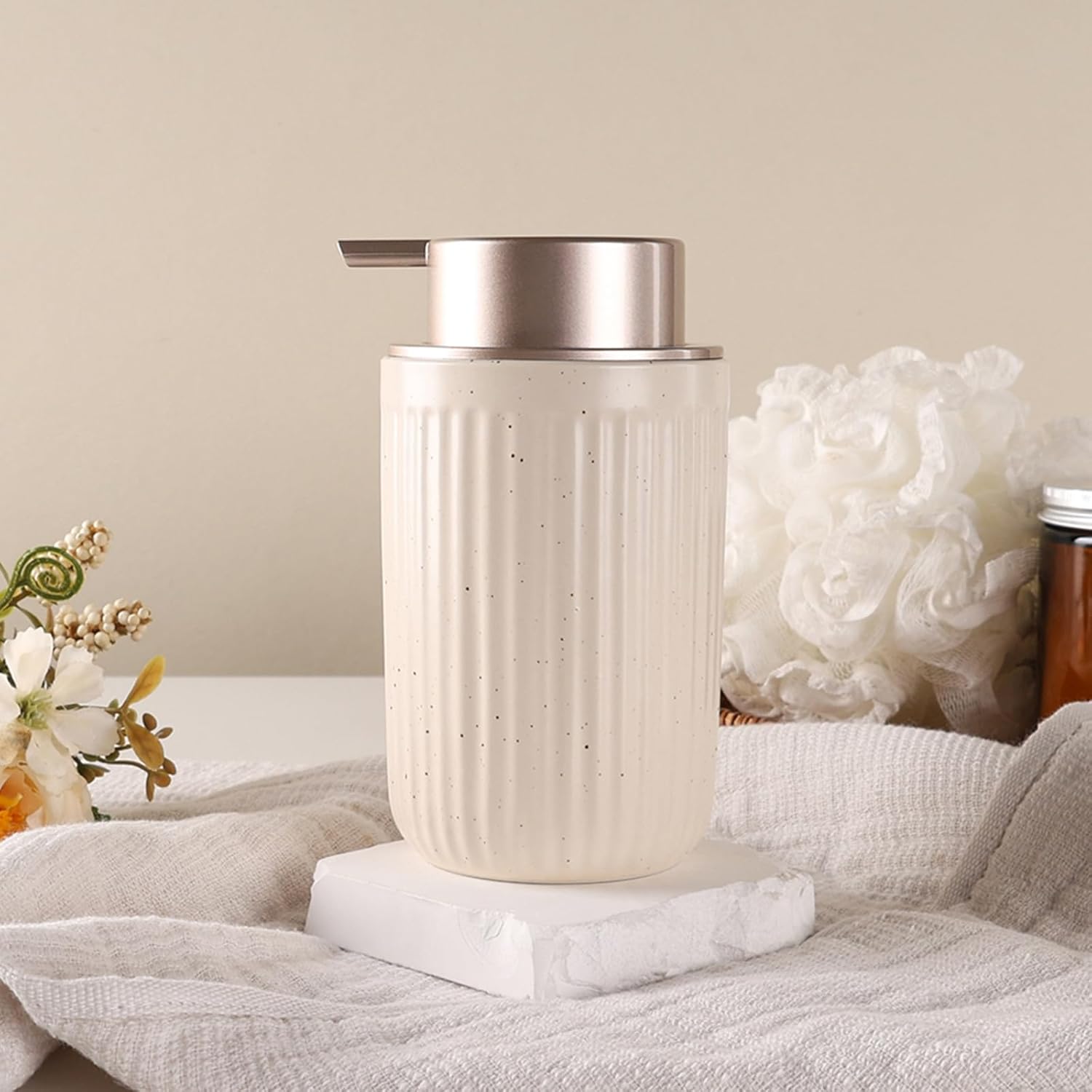 White soap dispenser on a textured surface with flowers and a candle in the background
