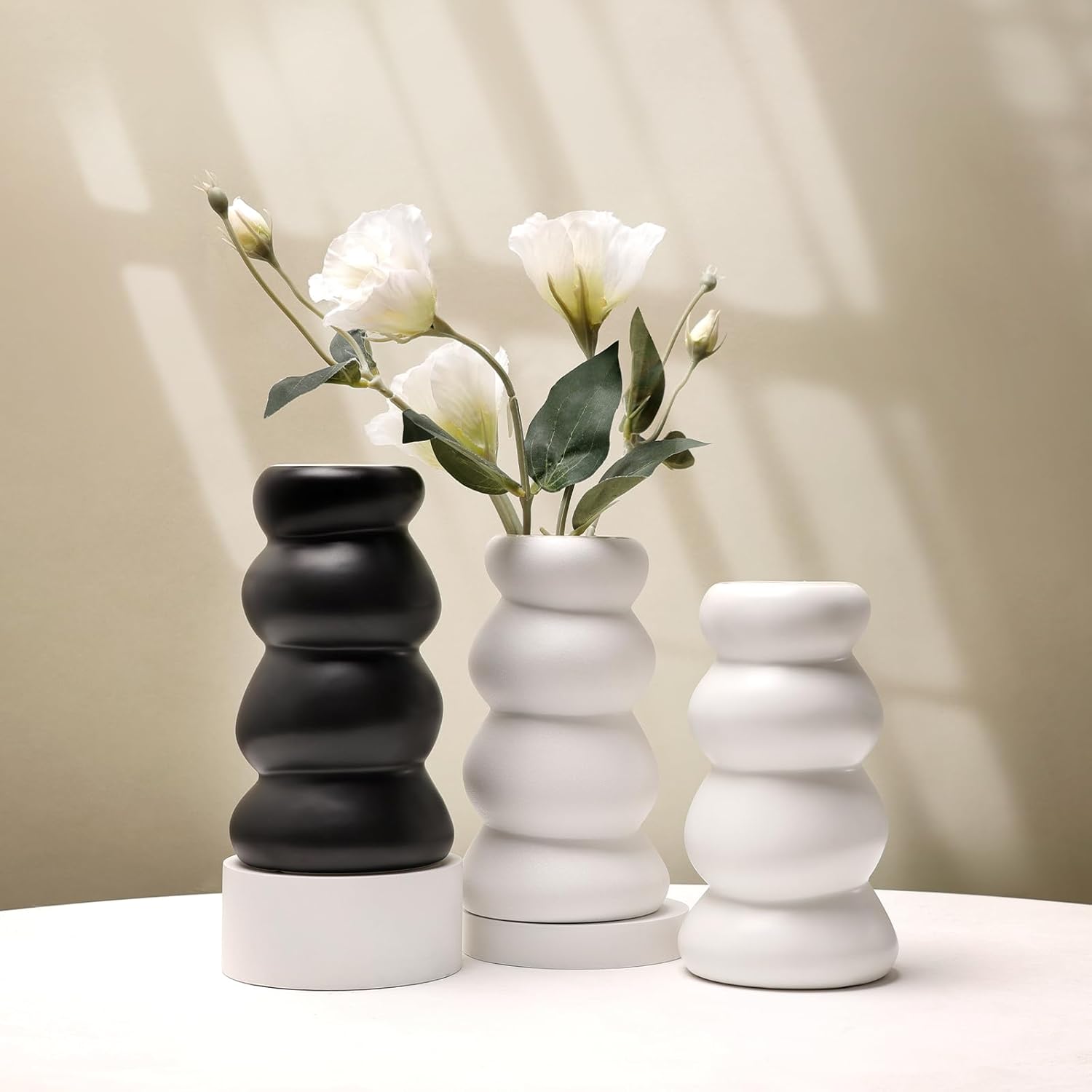 Three modern ceramic vases in black, white, and gray with a plant on a neutral background
