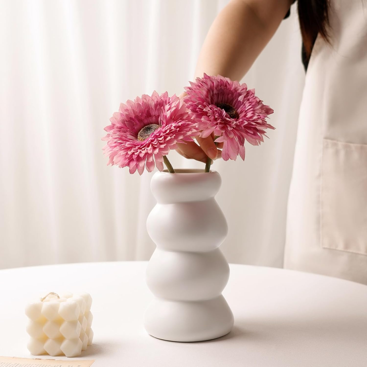 Lifestyle styling shot of the BosilunLife Stacked Bubble Flower Vase in matte white recycled eco-clay. A person is arranging two vibrant pink gerbera daisies into the sculptural, four-tier chubby ceramic vase. Styled on a bright white table next to a trendy white bubble cube candle with soft, sheer curtains in the background, creating a romantic, airy aesthetic.