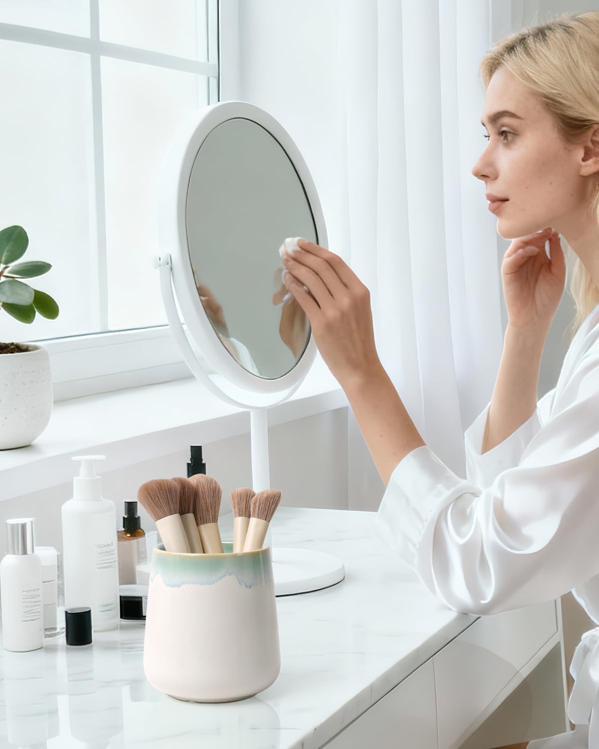 Lifestyle photograph of a BosilunLife eco-luxury ceramic tumbler used as a makeup brush holder. Crafted from GRS-certified recycled eco-clay, the vessel features a smooth white body with a soft sage green ombre rim. It is styled on a white marble dressing table next to a vanity mirror and skincare essentials. A woman in a white robe is seen in the background, illustrating a morning routine that blends Scandinavian functionality with the quiet balance of nature.
