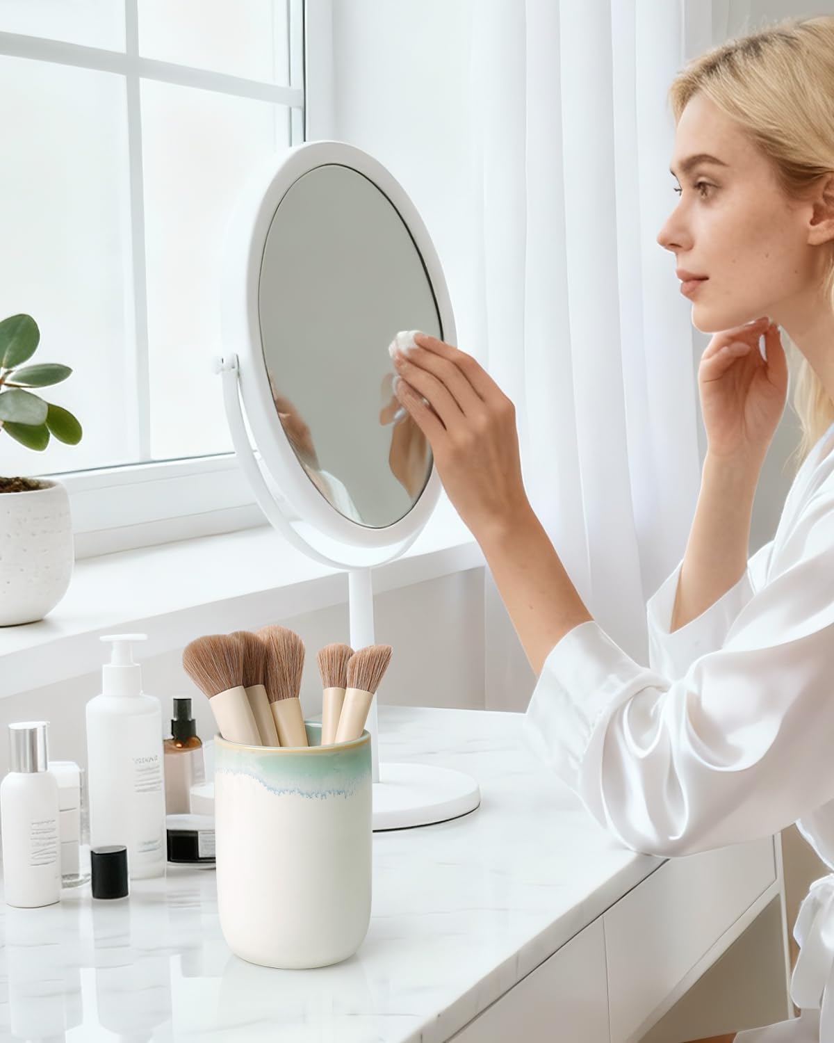 Lifestyle photograph of a BosilunLife eco-luxury ceramic tumbler used as a makeup brush holder. Crafted from GRS-certified recycled eco-clay, the vessel features a smooth white body with a soft sage green ombre rim. It is styled on a white marble dressing table next to a vanity mirror and skincare essentials. A woman in a white robe is seen in the background, illustrating a morning routine that blends Scandinavian functionality with the quiet balance of nature.