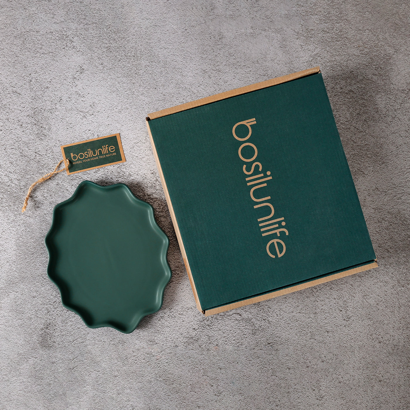 A flat-lay photograph showcasing a BosilunLife Forest Biome dinner plate in matte deep green beside its premium forest green branded gift box on a grey textured stone surface. The recycled eco-clay plate features a unique, organic scalloped wavy rim. The sustainable cardboard packaging is detailed with gold branding and a matching green brand tag with a rustic twine string, illustrating a refined, gift-ready unboxing experience for eco-friendly tableware.