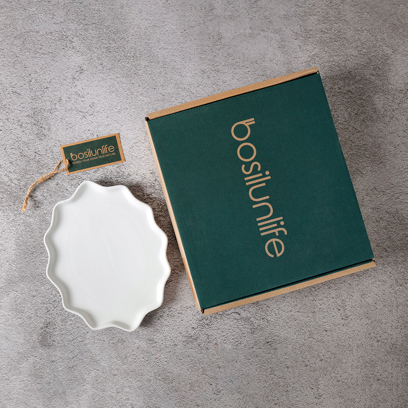 A flat-lay photograph showcasing a BosilunLife white matte Forest Star dinner plate beside its premium forest green branded gift box on a grey textured stone surface. The recycled eco-clay plate features a unique scalloped wavy rim. The sustainable cardboard packaging is detailed with gold branding and a matching green brand tag with a twine string, illustrating a refined, gift-ready unboxing experience for eco-conscious tableware.