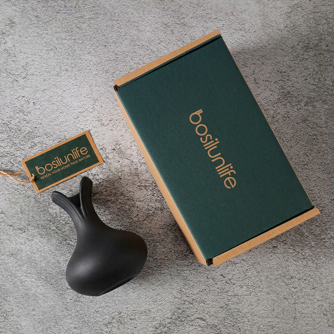 Flat lay of the BosilunLife sculptural black flower vase next to its premium sustainable gift packaging. The matte black recycled eco-clay vase rests on a textured grey concrete surface alongside a forest green and kraft cardboard box and a branded tag, highlighting an eco-luxury unboxing experience.