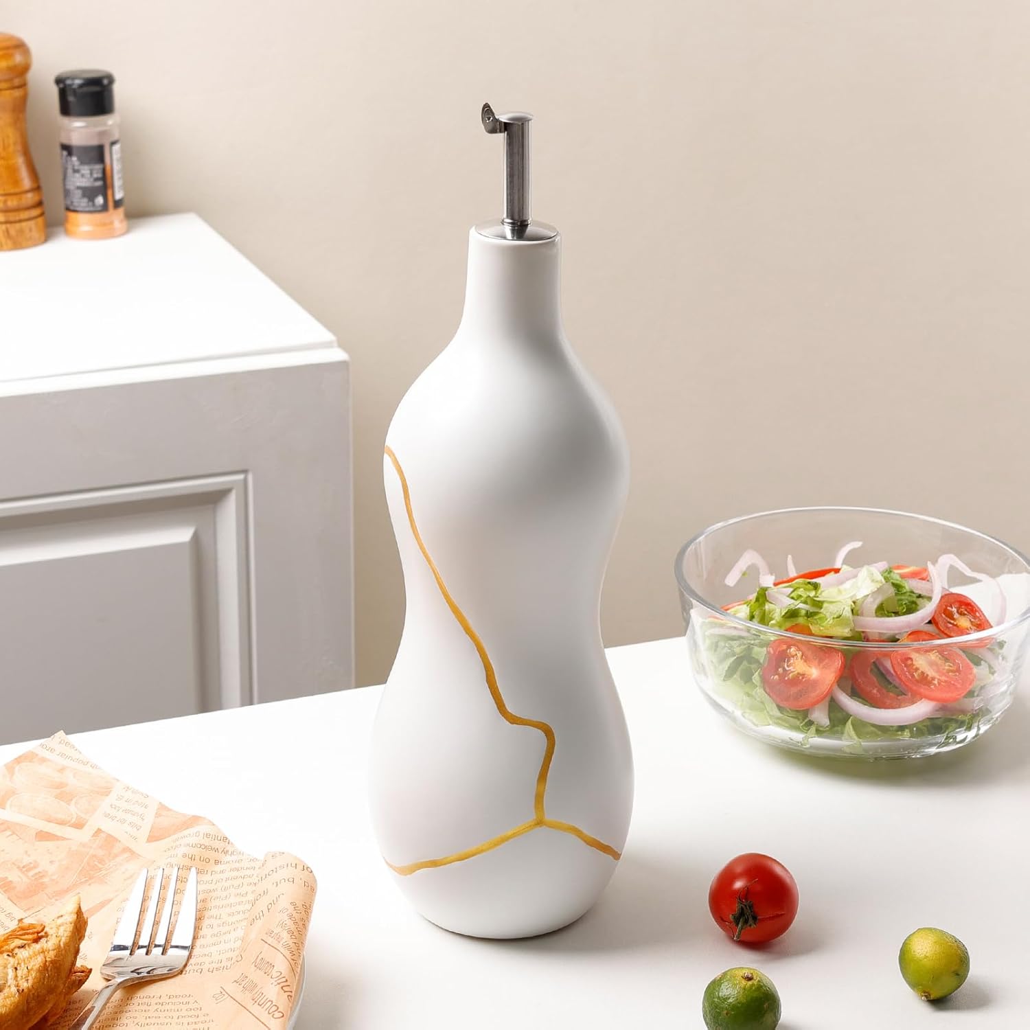 White ceramic oil dispenser on a kitchen counter with a salad and vegetables in the background.