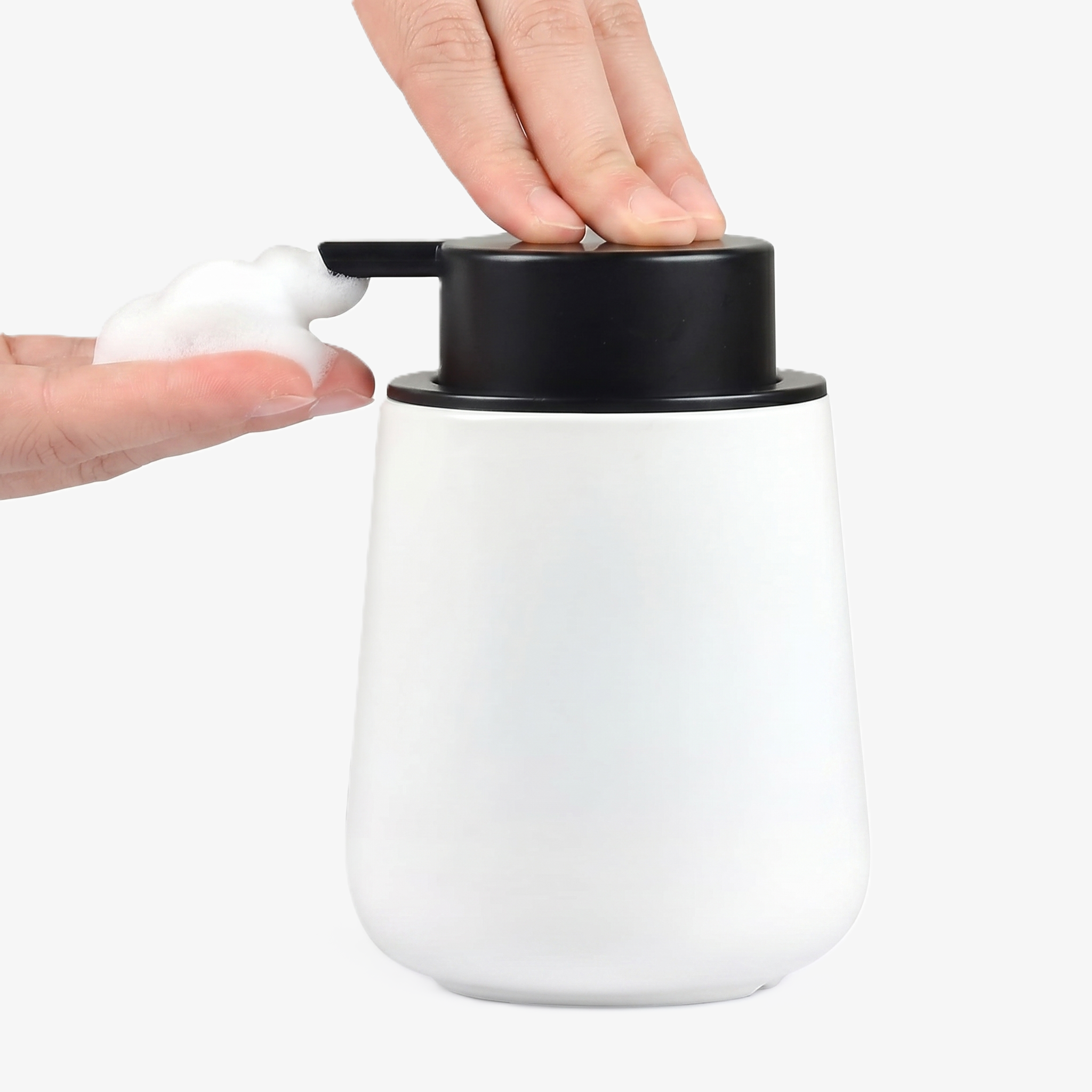 A person's hands operating the BosilunLife 12 oz eco-luxury foaming soap dispenser in a smooth matte white finish with a contrasting black pump. One hand presses the wide pump while the other catches a rich, velvety white foam. Crafted from GRS-certified recycled ceramic eco-clay, this organic rounded vessel reflects Scandinavian functionality and a commitment to a water-saving circular economy, providing a sophisticated organisation solution for a modern, minimalist washroom.