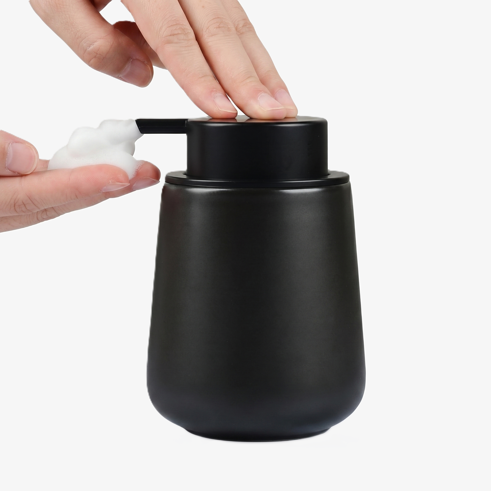 A person's hands operating the BosilunLife 12 oz eco-luxury foaming soap dispenser in a sleek matte black finish. One hand presses the wide matte black pump while the other catches a rich, velvety white foam. Crafted from GRS-certified recycled ceramic eco-clay, this organic rounded vessel embodies Scandinavian functionality and a water-saving circular economy, providing a sophisticated organisation solution for modern, minimalist washrooms.