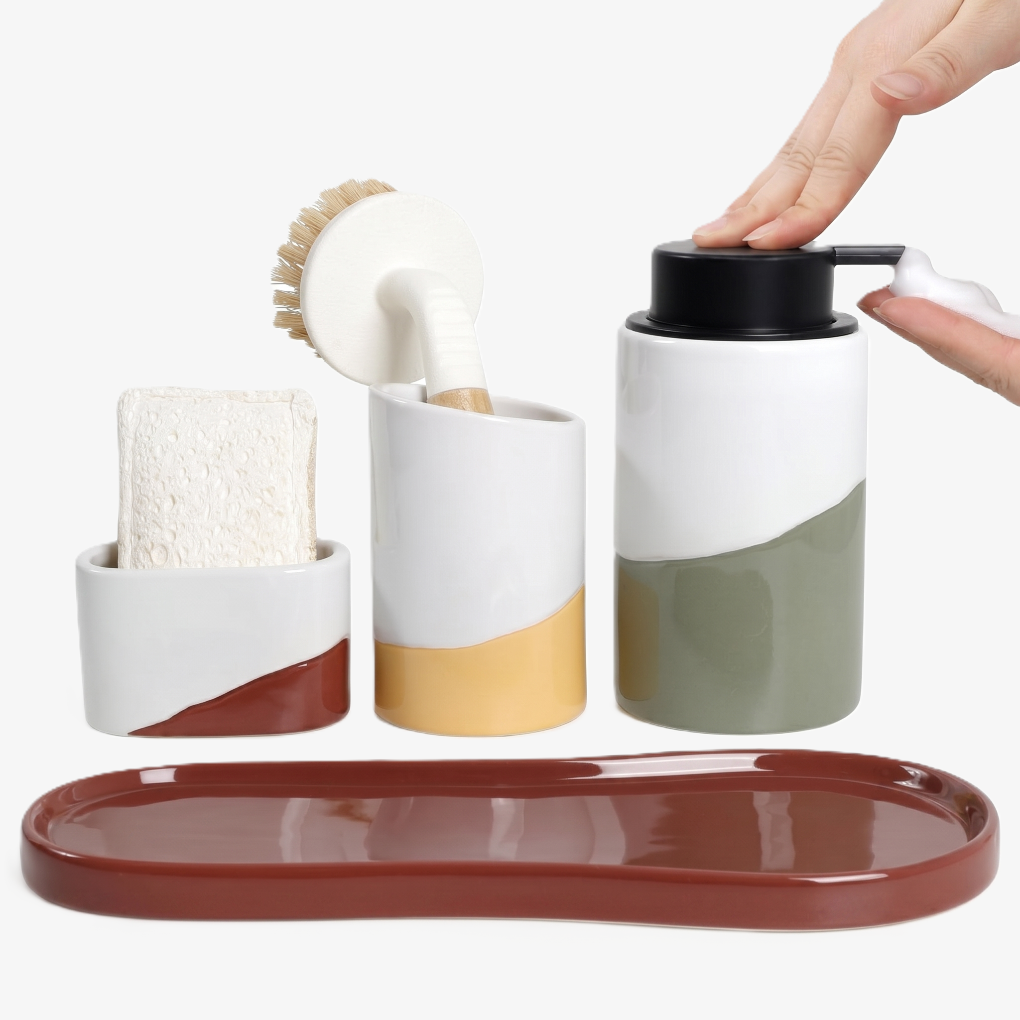 BosilunLife Waves 4-piece kitchen sink caddy set in recycled eco-clay. The vibrant collection features a terracotta tray, a white and terracotta sponge holder containing a natural sponge, a white and mustard yellow brush holder with a wooden dish brush, and a white and sage green foaming soap dispenser. A person's hands press the manual black pump to dispense clear dish soap, highlighting colourful and functional kitchen sink organisation.