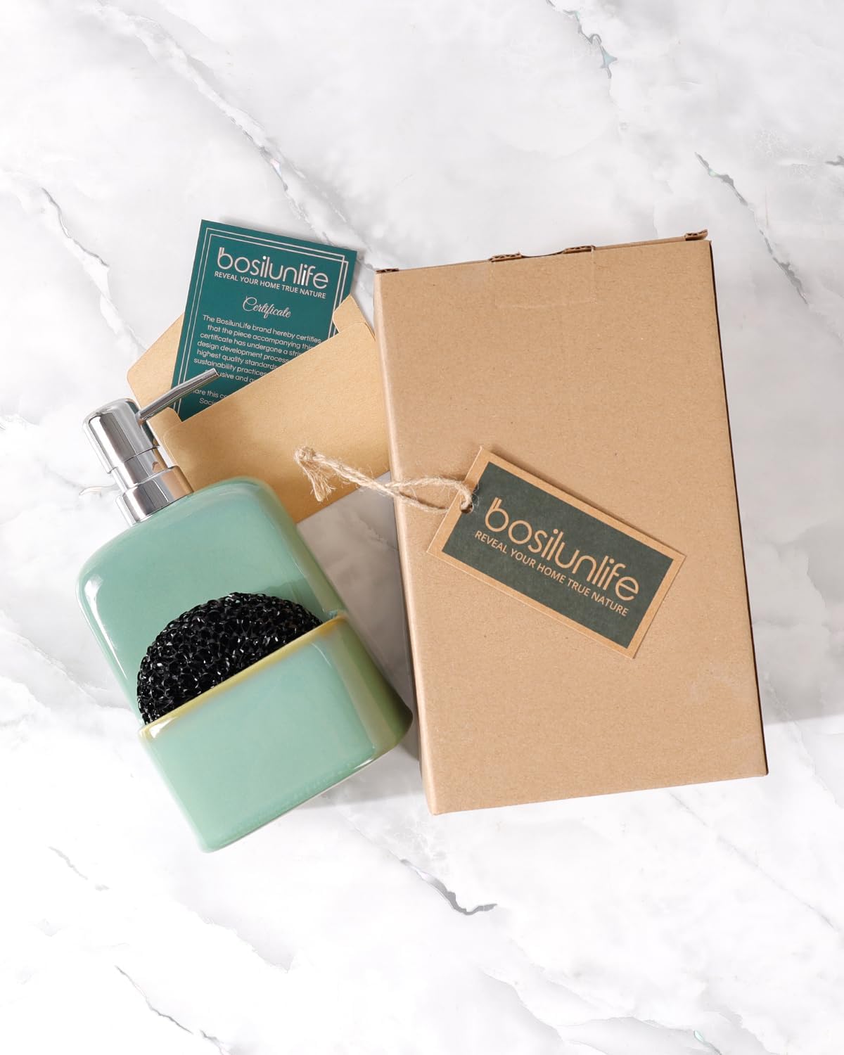 A glossy sage green BosilunLife 2-in-1 kitchen soap dispenser and sponge caddy made from recycled eco-clay. The minimalist ceramic dispenser is shown on a white marble surface next to its eco-friendly cardboard gift box, a green certificate of authenticity, and a branded tag with twine. A black mesh scouring pad is tucked into the integrated holder, illustrating a sustainable, gift-ready kitchen organisation solution.