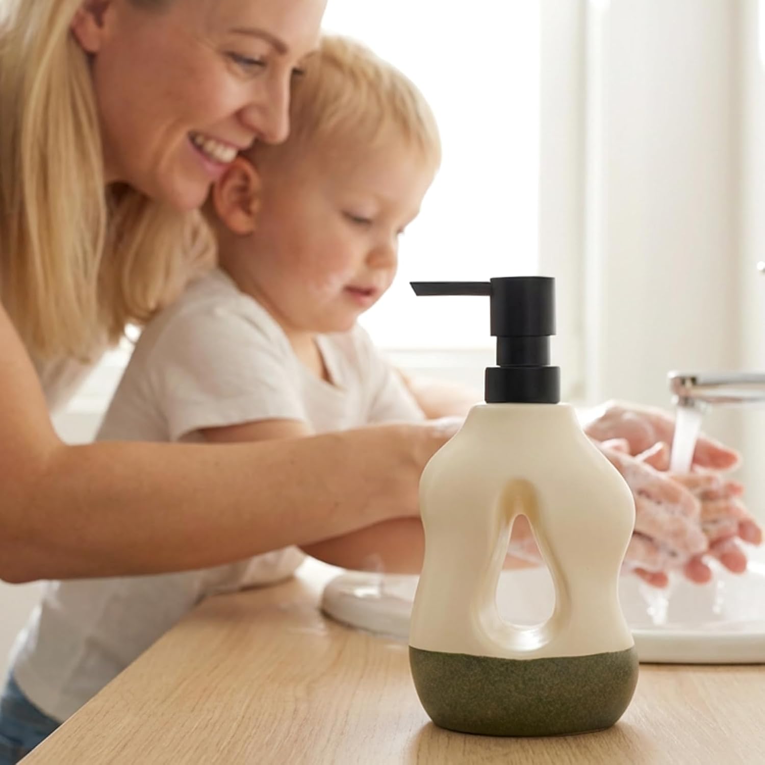 Environmental lifestyle shot of the BosilunLife Pebblebase liquid soap dispenser in moss green and ecru. The sculptural ceramic dispenser, crafted from recycled eco-clay, sits in sharp focus on a light wooden worktop. In the soft-focus background, a smiling woman helps a young child wash their hands at a sink, showcasing a non-toxic, sustainable, and family-friendly washroom aesthetic.