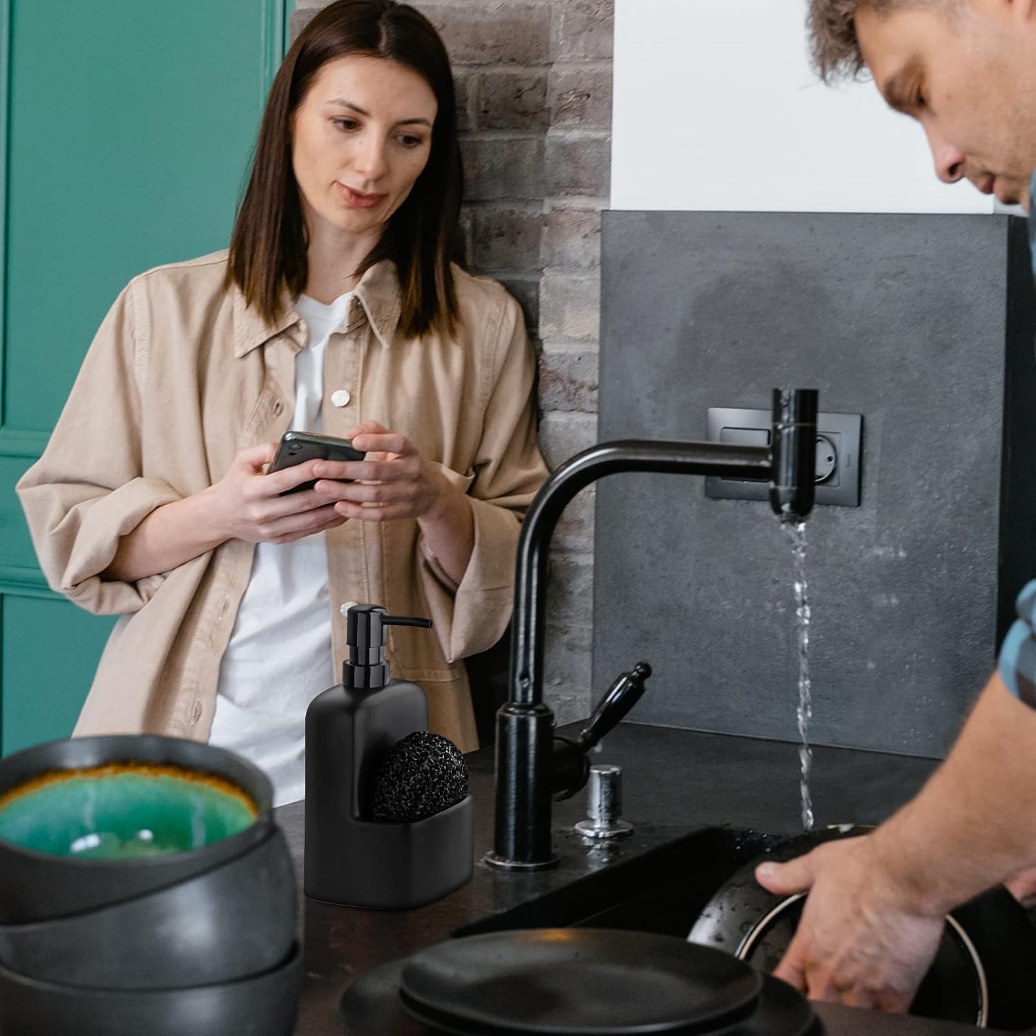 Person washing a pan under a modern black faucet in a kitchen, with another person using a phone.